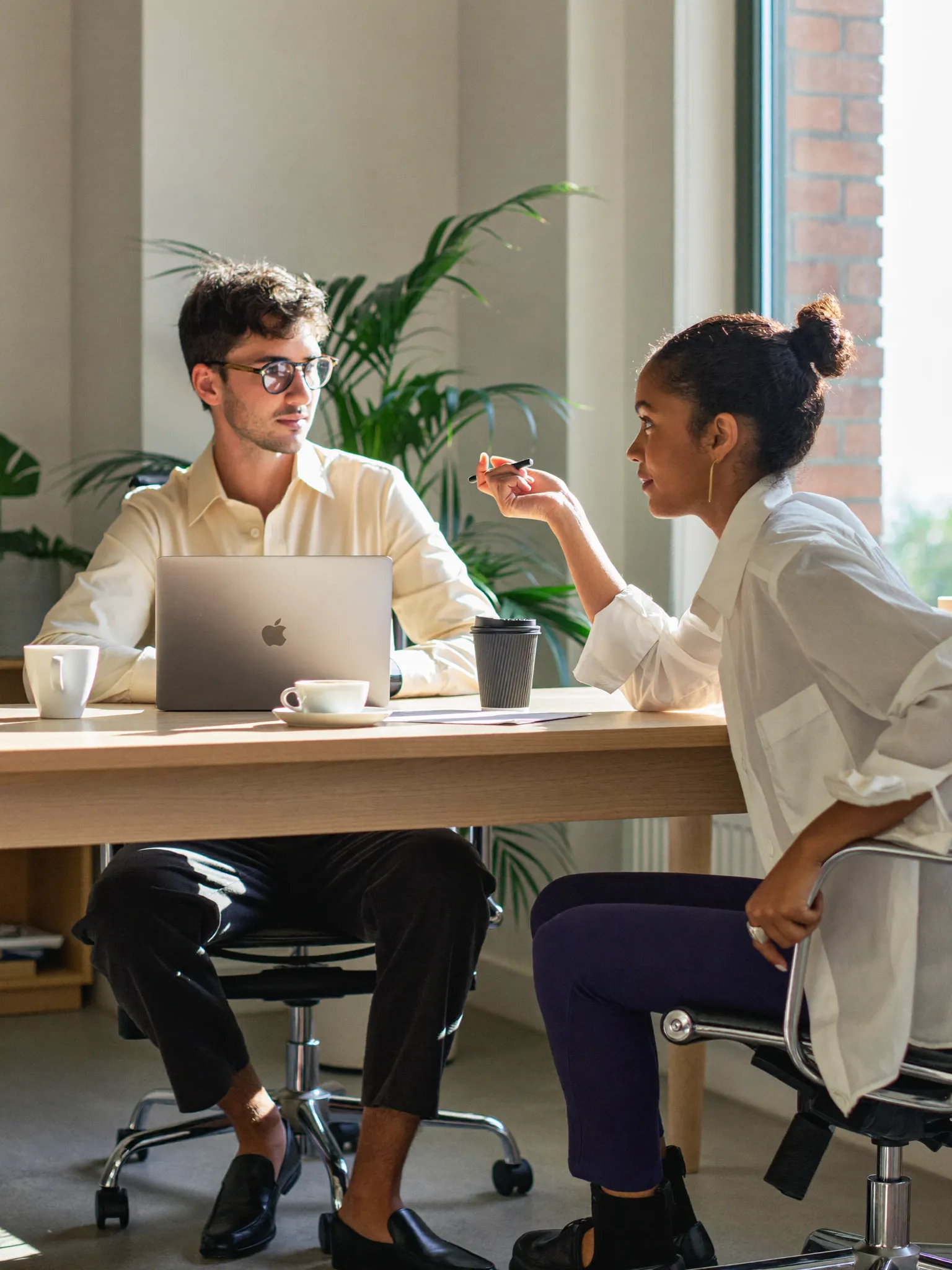Two colleagues in a relaxed one-to-one conversation at a desk in a bright, modern office